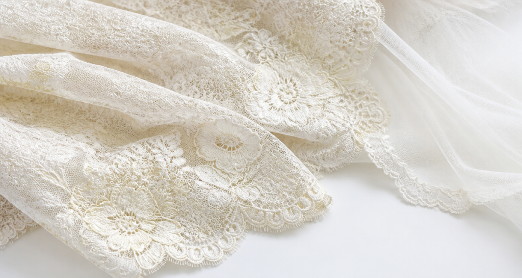 Layers of ivory bridal lace fabric with floral motifs draped beside a soft tulle veil on a white studio surface