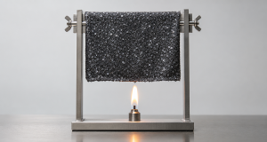A dark charcoal sequin fabric swatch held in a plain metal clamp frame above a small controlled teardrop flame in a laboratory flammability test, clean light-grey wall and stainless steel bench, fire safety compliance testing