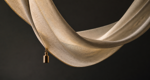 A flowing champagne-toned woven fabric draped in a sweeping curve against a dark charcoal background with a small polished gold weight hanging from its lower edge, emphasizing drape and motion