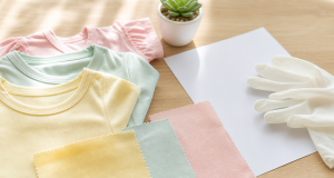 Pastel baby t-shirts in pale yellow, mint and pink with matching pinking-edged fabric swatches on a light wooden table beside white handling gloves, a blank white paper and a potted succulent in soft morning light