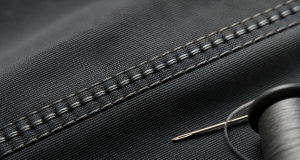 Extreme macro of a precise lock-stitch seam on charcoal fabric with a sewing needle and thread spool softly blurred in the corner