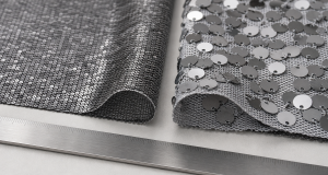 Two sequin fabric samples side by side on a light grey workbench: fine densely set small silver sequins on a dark tight-knit mesh base and larger loose silver paillettes on an open grey mesh, curled edges revealing the mesh structure, a brushed metal ruler at the lower edge