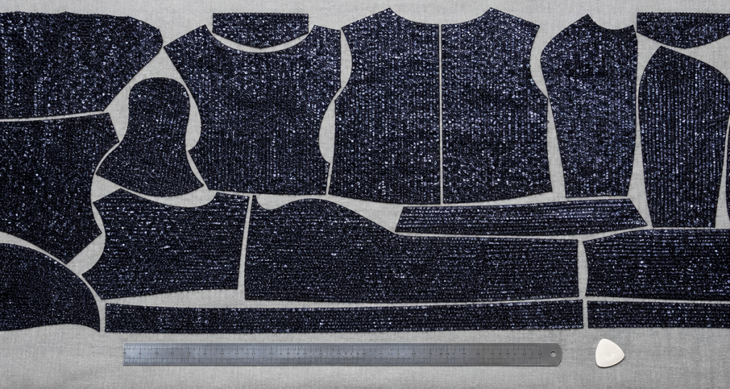 Top-down view of dark sequin garment pattern pieces tightly nested on a fabric cutting table with a steel ruler, minimal negative space in a precision cutting layout