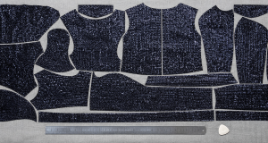 Top-down view of dark sequin garment pattern pieces tightly nested on a fabric cutting table with a steel ruler, minimal negative space in a precision cutting layout