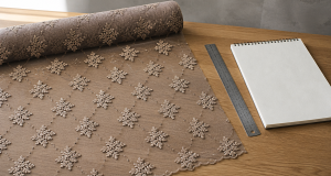 A roll of taupe embroidered lace mesh fabric unrolled across a light oak table showing a diagonal repeat of floral motifs with clean negative gaps between motifs, a stainless steel ruler and a blank spiral-bound sketchbook beside it, soft natural light