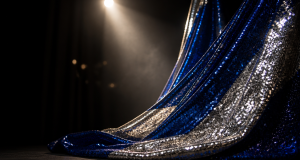 Cobalt blue and silver striped sequin fabric draped in deep folds under a bright stage spotlight with soft golden bokeh lights on a dark stage
