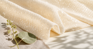 Soft ivory bio-based sequin fabric draped in waves with linen leaf and plant stem accents in bright natural daylight